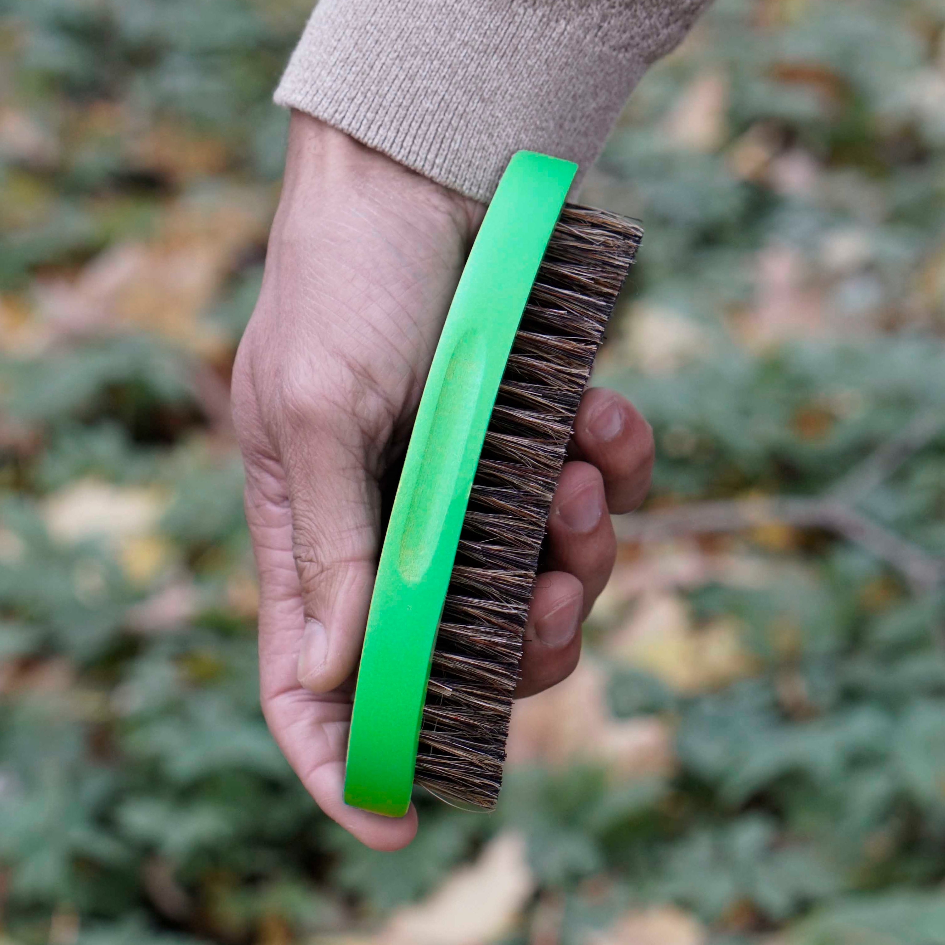 Hand holding a wave brush with a green handle against a blurred natural background